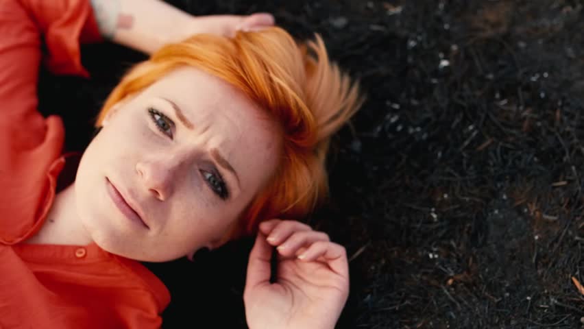 The sad girl with red hair lying on the black earth, sadly staring at the sky. Concept: need help, loneliness
