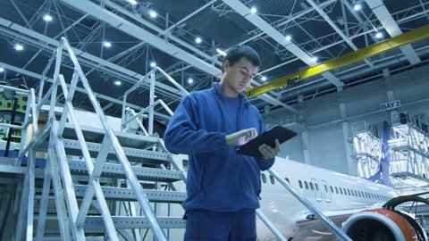 Aircraft maintenance mechanic is going down the stairs while using tablet and greeting his colleague a the bottom in a hangar. Shot on RED Cinema Camera in 4K (UHD).