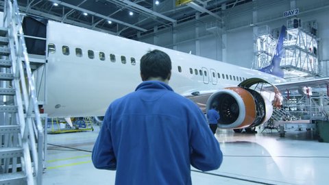 Aircraft maintenance mechanic in blue uniform is walking to greet his colleague who is checking the plane turbine blades in a hangar. Shot on RED Cinema Camera in 4K (UHD).