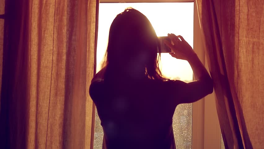 Young Girl Taking Photos Of City With blurred architectures on Mobile Smart Phone at home window in slow motion