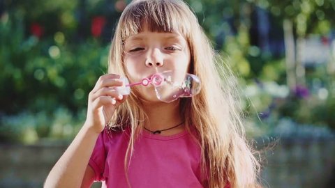 Cute little girl is blowing soap bubbles in the garden outdoors on a sunny day. Slow Motion 240 fps. Happy childhood concept. Child is playing in bright sun light. 