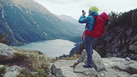 Hiker woman taking picture with smart phone from mountain peak with lake view. Tourist using photo app, touching screen. Stabilized Slow Motion 120 fps. Epic Steadicam shot. Misty Mountains Series. 