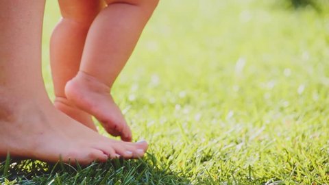 Little baby learns to walk. Close up on feet. Slow Motion 240 fps. Mother is teaching her child to do the first steps on a green grass in summer. Toddler is learning to walk outdoors on a green lawn.