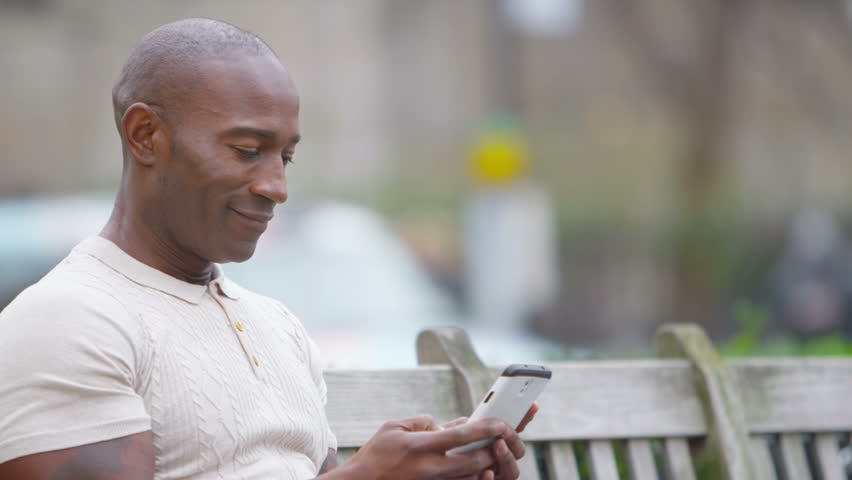 4K Portrait of casual smiling man sitting on park bench with mobile phone. Shot on RED Epic.