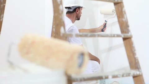 painter man at work, with roller painting wall, and wooden ladder in close up