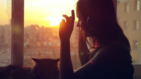 Young woman wears headphones and relaxing with her lovely Maine Coon cat at window with blurred city background at sunset in slowmotion. 1920x1080