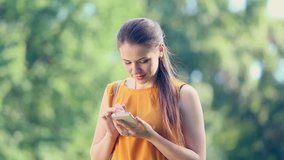 Young girl using smartphone outside at green trees nature background. Young woman on cell phone.  Woman texting sms on her phone outside in park.  Real time full hd video footage.