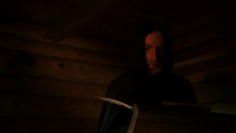 KIEV/UKRAINE - JUL 14 2015: Dark Room in an old log hut of the 11th century. Candlelight. Old man, monk, chronicler, dressed in black robe, in the hood. Nestor the Chronicler. He writes with a quill