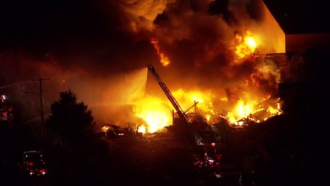 Night time aerial shot of a large building or warehouse fire. Fire department and emergency crews on scene to fight and extinguish the flames before the building is destroyed from wildfire spread. NX