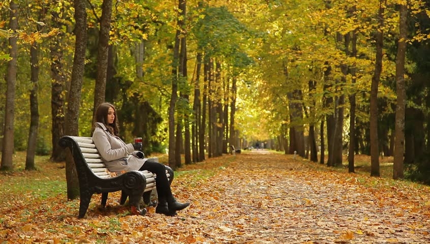 Young beautiful woman with thermos cup sitting on a bench in a beautiful autumn park.
