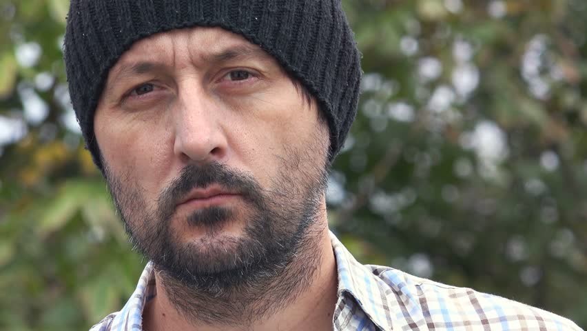 Serious confident lumberjack, adult bearded man holding a big axe and looking at camera in outdoors surrounding, close up of face, treetop in background.