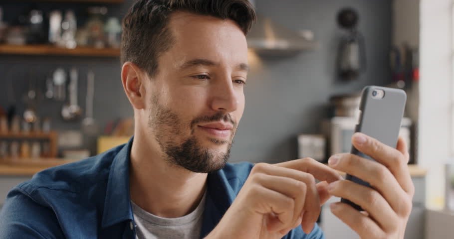 Attractive man at home using smartphone in kitchen sending message on social media smiling enjoying modern lifestyle wearing blue shirt Royalty-Free Stock Footage #12266195