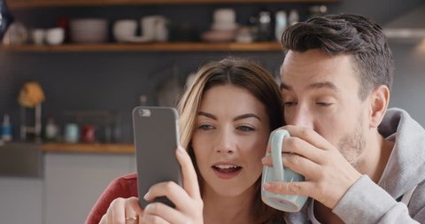 Happy couple at home in kitchen at breakfast using smartphone together browsing online having fun drinking coffee