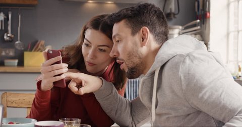Happy couple at home in kitchen at breakfast using smartphone together browsing online