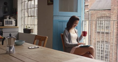 Beautiful Mixed race woman at home drinking coffee using digital tablet happy loft apartment wearing pajamas looking out of window