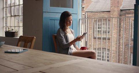 Beautiful Mixed race woman at home drinking coffee using smartphone loft apartment wearing pajamas