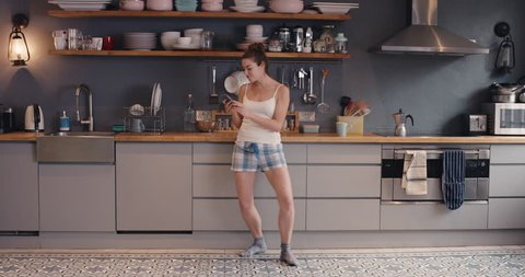 Happy young woman dancing in kitchen wearing pajamas in the morning listening to music on smartphone and coffee at home