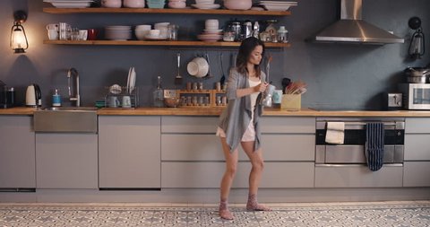 Happy young woman dancing in kitchen wearing pajamas in the morning listening to music on smartphone and coffee at home