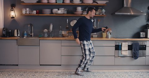 Happy young man dancing in kitchen wearing pajamas in the morning listening to music on smartphone and coffee at home
