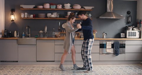 Happy young couple newly wed dancing listening to music in kitchen wearing pajamas coffee morning at home having fun