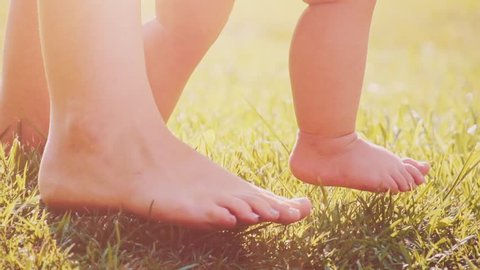 Little baby learns to walk. Close up on feet. Slow Motion 240 fps. Mother is teaching her child to do the first steps on a green grass in summer. Toddler is learning to walk outdoors on a green lawn.