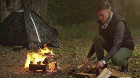 Short haired man in autumn clothes is chopping wood with an axe and throwing it into the fire. Shot on RED Cinema Camera in 4K (UHD).の動画素材