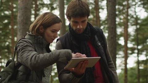 Long haired man and brunette woman in dark autumn clothes are standing in a forest and using tablet. Shot on RED Cinema Camera in 4K (UHD).