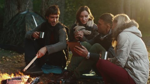 Group of men and women sit next to a campfire with drinks and use tablet in a forest. Shot on RED Cinema Camera in 4K (UHD).