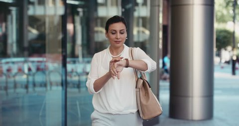 Smart Middle Eastern businesswoman using smartwatch commuting to work entering glass corporate building smiling