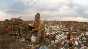 man  homeless beggar sitting in a landfill with a hat asks for money food waste