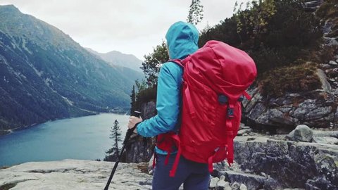 Hiker woman walking with backpack admiring mountain lake view landscape from the cliff edge. Stabilized, Slow Motion 120 fps. Epic Steadicam hiking in a stormy wind. Misty Mountains Series.
