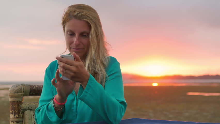 young woman sitting outdoor holding and lookin at smartphone enjoying vacation surfing on internet sea sunset background