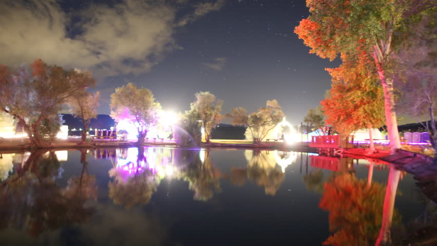 Motion controlled astrophotography time lapse footage with pan left & tilt up motion of starry sky over illuminated trees surrounding reflective pond at Joshua Tree Music Festival 2015 -Long Shot-