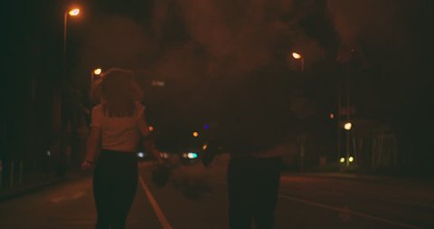 Teenage guy and girl running down a late night city street carrying smoke flares under the street lights in Slow Motion