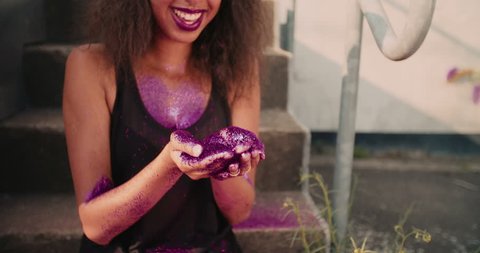 Gorgeous Afro American girl wearing a cap over her soft curly hair blowing bright, pink coloured, sparkling glitter from her hands while outdoors in Slow Motion Video stock