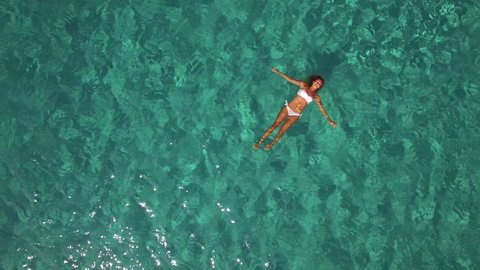 Aerial - Beautiful young caucasian woman with fit body shape in white bikini floating on water surface in crystal clear turquoise ocean