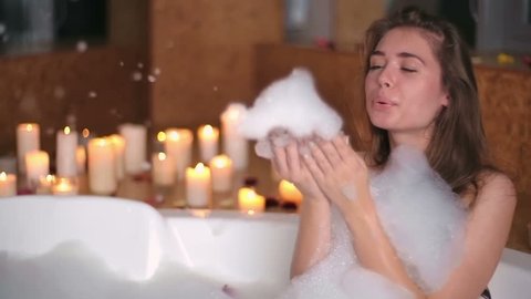 Attractive young woman blowing soap foam in a bath surrounded by burning candles 