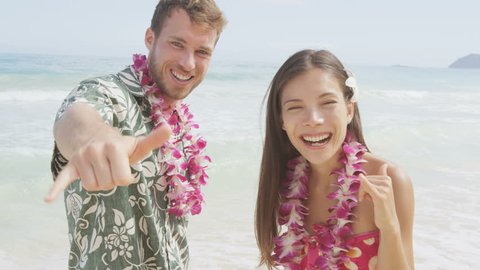 Happy couple of tourists standing on Hawaiian beach at their Hawaii vacation. Asian woman and Caucasian man wearing flower lei garland and Aloha clothing showing Shaka hand sign on travel. RED EPIC.