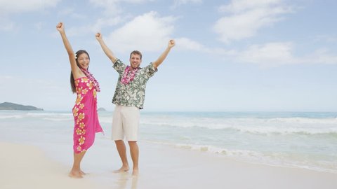 Celebrating cheering happy winning couple on Hawaii beach beach on Hawaiian vacation. Asian woman and Caucasian man wearing flower lei garland and Aloha clothing raising arms up dancing of joy.