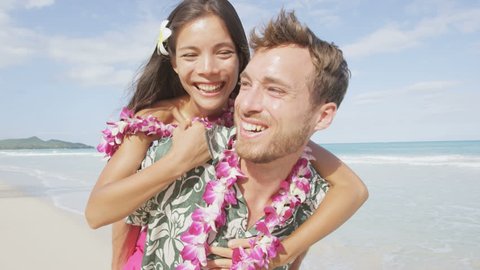 Beach couple having fun laughing on Hawaii holiday. Beautiful Asian mixed race woman piggybacking on Caucasian boyfriend wearing traditional Hawaiian lei on Oahu, Hawaii, USA. RED EPIC SLOW MOTION.