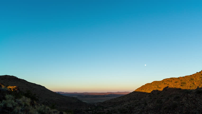 A static timelapse tilting up towards a bright blue sky at sunrise over vast mountain landscape with wide open plains below as the moon sets. 4K