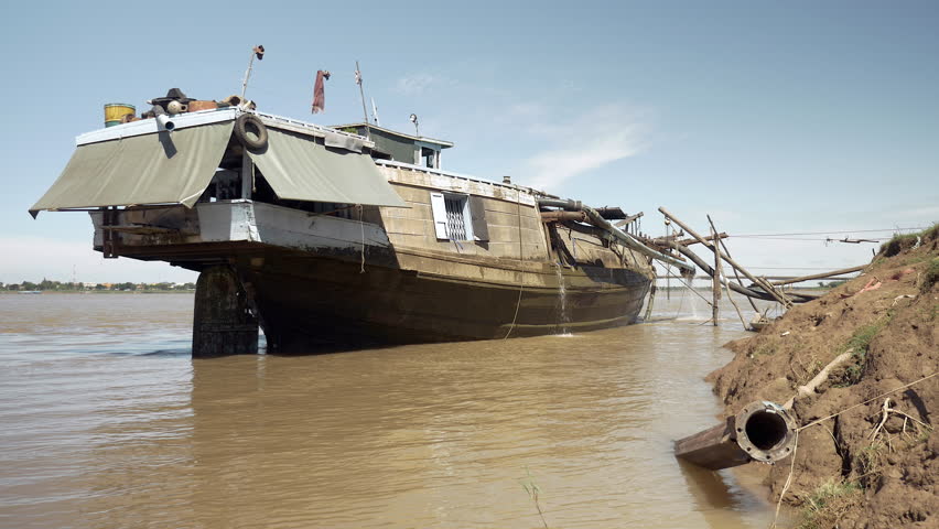  Side view of a dredging boat tied to shore; Sand is being pumped out of the boat through a system of pipes hold between bamboo canes