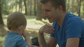 Father helps crying son to use cell phone in park.