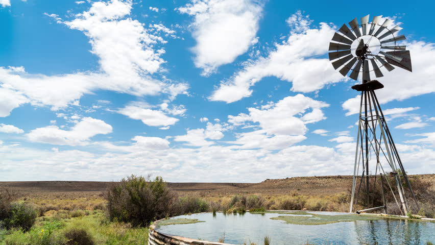 A linear midday timelapse of a windmill blowing in the wind next to and old zinc farm dam with scattered clouds against a bright blue sky in a typical Karoo landscape. 4K