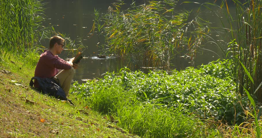 Man in Glasses is Sitting on The Sloping Lake Bank, Reading the Book with Brown Cover, Leafing through the Pages, Reader, Backpack, Man Has Closed the Book, Put it Into Backpack, forest lake, wild