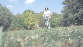 Woman joyfully riding bike through a green grass glade over forest / slowmotion / video from the ground