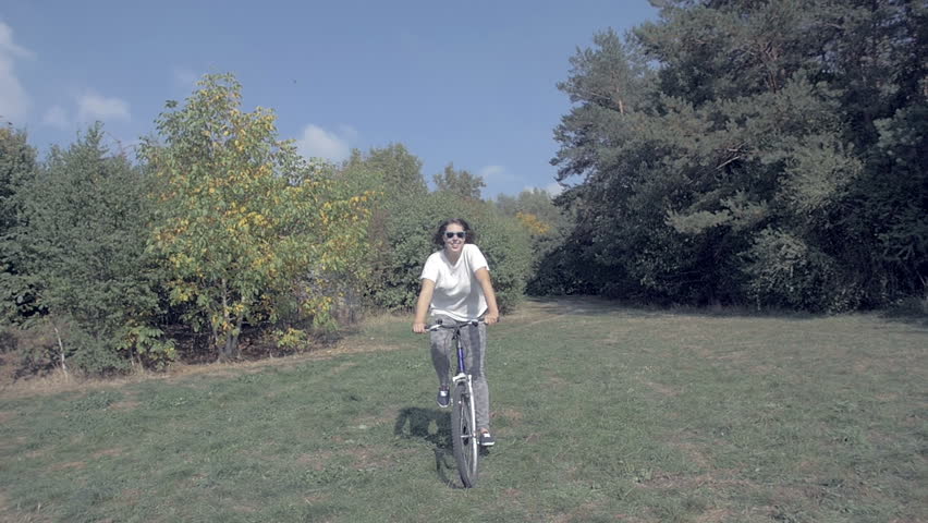 Woman smiling and joyfully riding bike through a green grass glade over forest / slowmotion