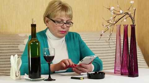 Senior woman sits at the table, drinks red wine and uses on smartphone
