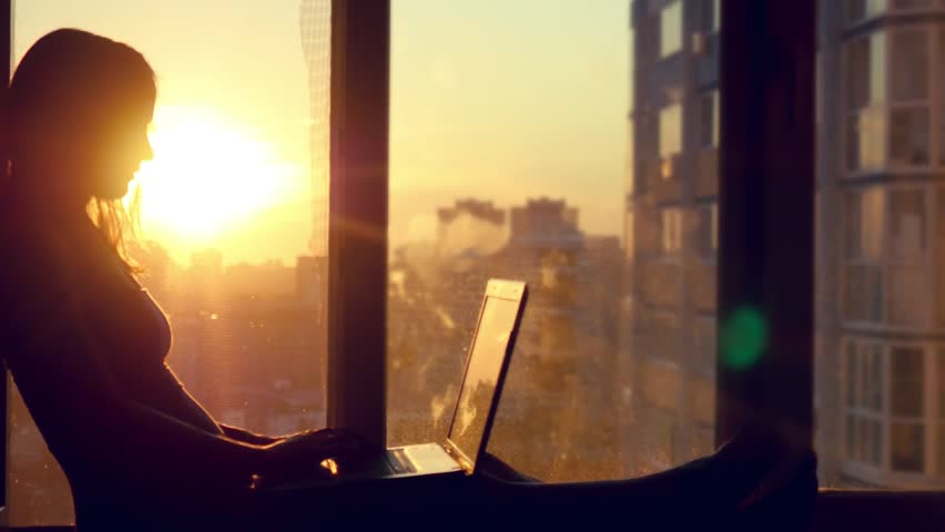 Beautiful young brunette business woman working on her laptop. Heaven sunset glass background behind her. Girl sitting on the window on modern building blurred background in city | Shutterstock HD Video #12406052