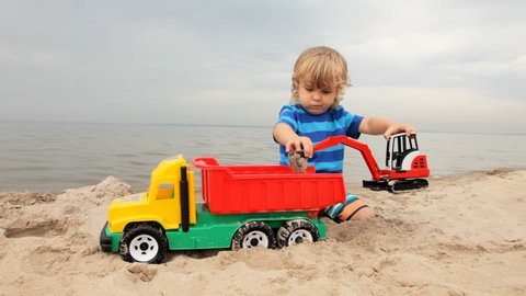 little child boy playing with toy excavator and big truck on the sand beach